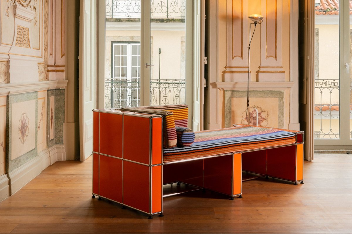 Diván naranja de estructura metálica con tapizado de rayas de colores, junto a una lámpara de pie, en una sala con grandes ventanales