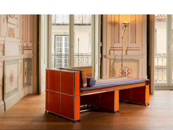 Diván naranja de estructura metálica con tapizado de rayas de colores, junto a una lámpara de pie, en una sala con grandes ventanales